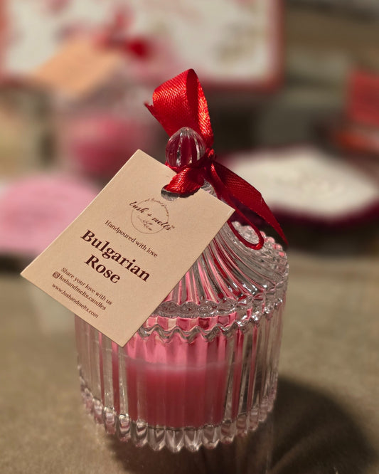 Pink candle in a glass container with a red ribbon and tag labeled 'Bulgarian Rose'.